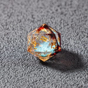 May include: A clear, multi-faceted D20 die with gold numbering. The die has red and gold accents, with a blue, glittery core. The die is designed for tabletop role-playing games.