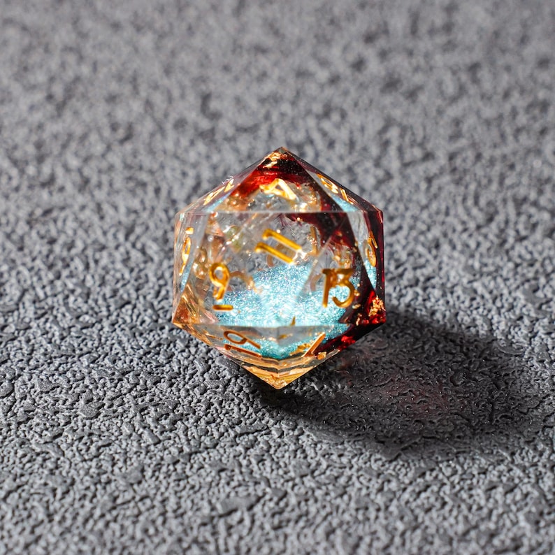 May include: A clear, multi-faceted D20 die with gold numbering. The die has red and gold accents, with a blue, glittery core. The die is designed for tabletop role-playing games.