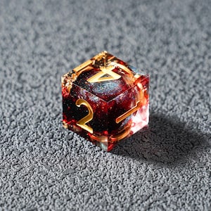 May include: A clear, six-sided die with gold numbering and a red and gold interior. The die has a glittery, celestial-like core and is set against a textured gray surface. The numbers are in a gold, serif font.