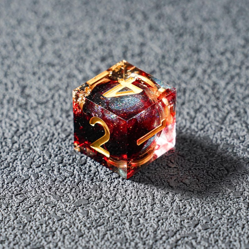 May include: A clear, six-sided die with gold numbering and a red and gold interior. The die has a glittery, celestial-like core and is set against a textured gray surface. The numbers are in a gold, serif font.