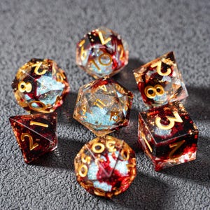 May include: A set of seven polyhedral dice for tabletop games. The dice are clear with red and gold accents, and gold numbering. Some dice have a blue glitter effect inside. The dice are arranged on a gray surface.