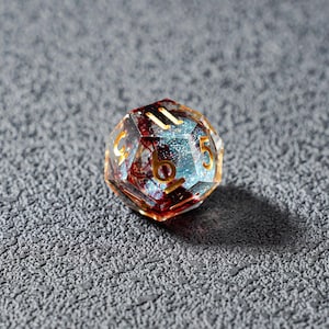 May include: A multi-faceted, clear resin die with gold numbering. The interior features a mix of red and blue inclusions, with a glittery, light blue core. The die is set against a textured, gray surface.