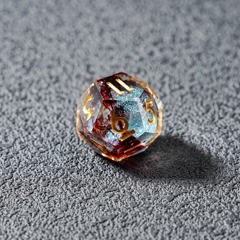 May include: A multi-faceted, clear resin die with gold numbering. The interior features a mix of red and blue inclusions, with a glittery, light blue core. The die is set against a textured, gray surface.