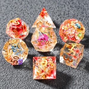Könnte beinhalten: Set aus sieben polyedrischen Würfeln für Tabletop-Spiele. Jeder Würfel ist transparent mit eingebetteten bunten Blumen und goldfarbenen Zahlen. Die Würfel umfassen einen d4, d6, d8, d10, d12, d20 und einen Prozentwürfel.