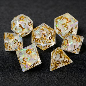Puede incluir: Un juego de siete dados poliédricos hechos de resina transparente con escamas iridiscentes y brillantes incrustadas. Cada dado presenta números grabados con un acabado dorado metálico, incluyendo un d20, d12, d10, d8, d6, d4 y un dado de porcentaje.