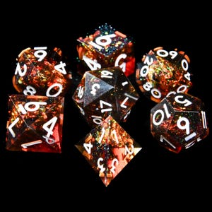 以下が含まれることがあります： 暗い琥珀色の樹脂ベースにカラフルなグリッターが埋め込まれた、7つの多面体サイコロのセット。サイコロには白い数字が書かれており、黒い面に配置されています。セットには、D4、D6、D8、D10、D12、D20、およびパーセンタイルダイスが含まれています。