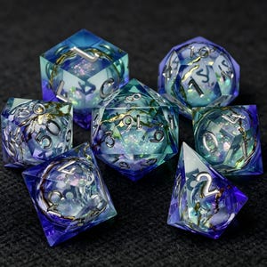 Könnte beinhalten: Ein Set aus sieben polyedrischen Würfeln aus durchscheinendem blauem und violettem Harz. Jeder Würfel enthält einen kugelförmigen Kern mit irisierendem Glitzer und goldfarbenen Metalleinschlüssen. Die Zahlen sind in Silber eingraviert.