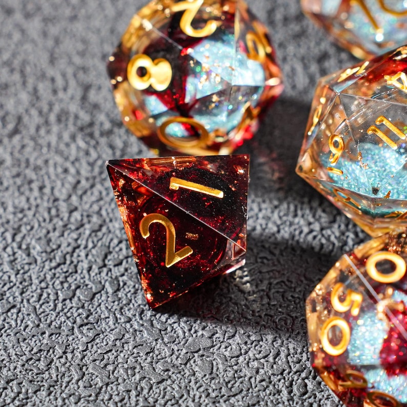 May include: Close-up of several polyhedral dice. The dice are a mix of red, gold, and blue, with gold numbering. The dice are on a textured gray surface. The dice are for tabletop games.