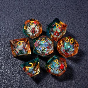 Puede incluir: Juego de siete dados poliédricos con diseño floral. Los dados son transparentes con tonos naranja, azul y verde. Los números son dorados. El diseño floral incluye flores rojas y blancas.