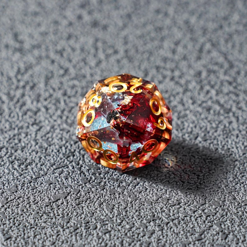 May include: A multi-faceted, twenty-sided die with a transparent body, filled with red and gold accents. The numbers are gold. The die is designed for tabletop role-playing games.