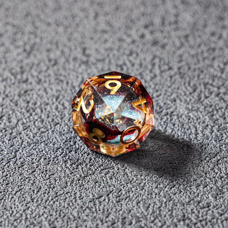 May include: A multi-faceted, clear resin D20 die with gold numbering. The interior features a mix of red, gold, and blue inclusions, creating a galaxy-like effect. The die is designed for tabletop role-playing games.