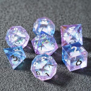 Può includere: Set di sette dadi poliedrici blu e viola. Ogni dado trasparente contiene glitter iridescenti e numeri bianchi. I dadi sono disposti su una superficie grigia.