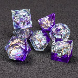Puede incluir: Juego de siete dados poliédricos de diferentes formas. Los dados son una mezcla de resina morada y transparente con detalles dorados y números blancos. Diseñados para juegos de rol de mesa.