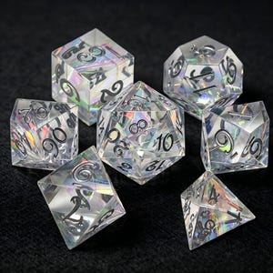Puede incluir: Un juego de siete dados poliédricos hechos de material transparente y claro con un efecto de lámina holográfica iridiscente en el interior. Cada dado presenta números negros profundamente grabados, incluyendo un d20, d12, d10, d8, d6, d4 y un dado de porcentaje.