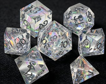 Puede incluir: Un juego de siete dados poliédricos hechos de material transparente y claro con un efecto de lámina holográfica iridiscente en el interior. Cada dado presenta números negros profundamente grabados, incluyendo un d20, d12, d10, d8, d6, d4 y un dado de porcentaje.