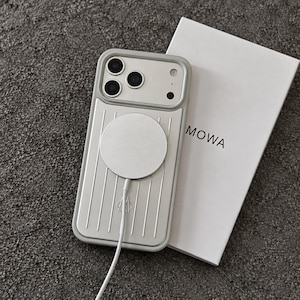 May include: A light gray phone case with a vertical grooved texture and a circular magnetic charger attached to the back. The case features a camera cutout and a small logo. Beside it lies a white rectangular box with the text RIMOWA printed on it.