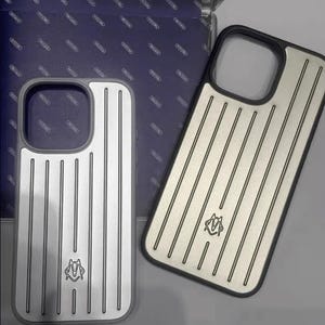 Op de afbeelding: Twee zilveren en zwarte telefoonhoesjes met verticale lijnen en een logo. De hoesjes zijn ontworpen om op een telefoon te passen, met uitsparingen voor de camera en knoppen. De hoesjes worden gepresenteerd op een blauwe doos.