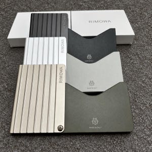 以下が含まれることがあります： 黒、銀、シャンパン色の溝付き外装を持つ3つのRimowaカードホルダー。各ホルダーの前面にはRimowaのロゴがあり、黒、グレー、またはオリーブ色の内部スリーブには「Made in Italy」というテキストとロゴが記されています。