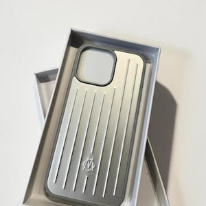 Peut inclure: Une coque de téléphone argentée et grise avec une texture nervurée verticale et un logo central, dotée d'une découpe carrée pour l'appareil photo. La coque est présentée dans une boîte rectangulaire blanche au design épuré et finition métallique.