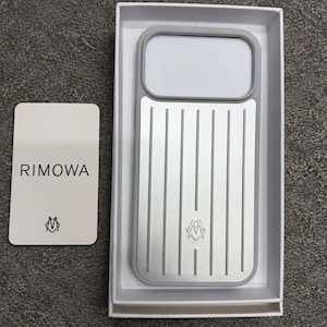 May include: A silver and grey phone case with a vertical grooved pattern and a small logo, presented in a white box alongside a white card featuring the text RIMOWA and a small logo. The case has a rectangular cutout at the top.