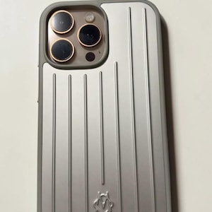 Puede incluir: Una funda para smartphone de color plateado con una textura de rayas verticales y un pequeño logotipo en relieve en la parte inferior central. La funda tiene un borde gris y un recorte para el sistema de triple cámara, con tres lentes circulares con bordes en oro rosa.
