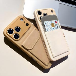 Könnte beinhalten: Zwei beige und cremefarbene Handyhüllen mit Kartenfächern. Eine Hülle ist geöffnet und zeigt eine Kreditkarte. Die andere Hülle hat eine Klappe, die die Kameralinsen abdeckt. Die Hüllen liegen auf einer weißen Oberfläche.