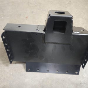 Pode incluir: Um suporte de metal preto com uma base retangular e uma estrutura vertical em forma de caixa. O suporte tem vários orifícios ao longo das bordas e uma abertura quadrada. Parece ser um componente para máquinas ou construção.