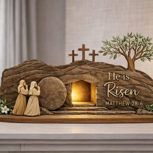 Può includere: Scultura religiosa marrone dettagliata raffigurante il sepolcro vuoto con due figure, tre croci e un albero. Il testo "È risorto" e Matteo 28:6 sono visibili. Fiori decorativi sono posti sulla base.