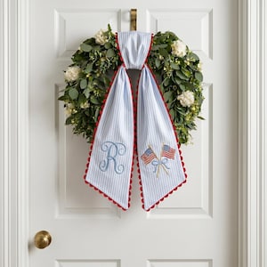 May include: A decorative wreath with a green foliage base and white flowers. A blue and white striped fabric with red scalloped edges hangs from the top, featuring an embroidered letter "R" and two American flags. The wreath is hanging on a white door.
