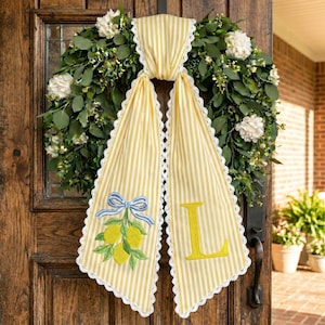 May include: A door wreath with a yellow and white striped fabric accent. The fabric has a lemon and bow embroidery and the letter "L". The wreath is made of greenery and white flowers.