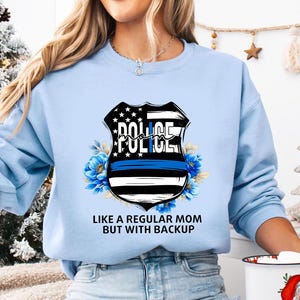 Peut inclure: Sweat-shirt bleu clair avec un motif d'écusson de police, une bande bleue et des motifs floraux. L'écusson porte l'inscription "POLICE" et le texte "LIKE A REGULAR MOM BUT WITH BACKUP" est imprimé en dessous.