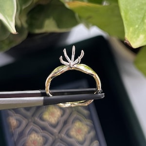 May include: A gold ring with a twisted band design featuring a silver-toned prong setting at the top. The ring is held by metal tweezers, showcasing the metallic texture and the contrast between the yellow gold band and the silver-coloured prongs.