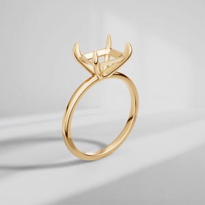 Puede incluir: Un engaste de anillo de oro amarillo pulido con una cabeza de forma cuadrada y cuatro garras diseñadas para sujetar una piedra preciosa. La banda es fina y circular, mostrando un acabado metálico liso y reflectante en toda la pieza.