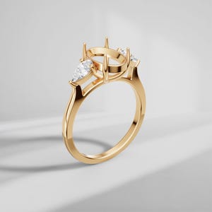Puede incluir: Un engaste de anillo en oro con un soporte central circular de garras para una piedra, flanqueado por dos piedras claras en forma de pera a cada lado. La banda es lisa y pulida, mostrando una configuración de tres piedras con engaste alto.