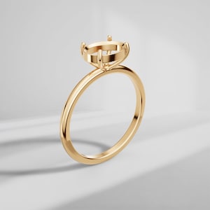 Puede incluir: Un engaste de anillo en oro pulido con una cabeza circular de cuatro garras diseñada para sostener una piedra preciosa. El aro es fino, liso y redondeado, mostrando un acabado metálico reflectante en toda la pieza de joyería.
