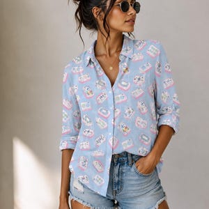 May include: A light blue button-down shirt featuring a repeating pattern of colorful mahjong tiles. The shirt has a classic collar, long sleeves with rolled cuffs, and a button-front closure. The fabric displays various tile designs in pink, white, and green.