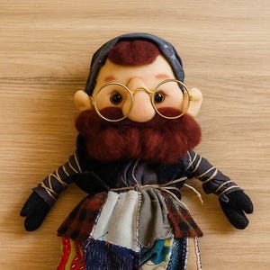 May include: A handmade fabric doll with a whimsical design. The doll features a burgundy beard, round gold glasses, and a patchwork outfit in various colors and patterns. The doll's arms and legs are black.