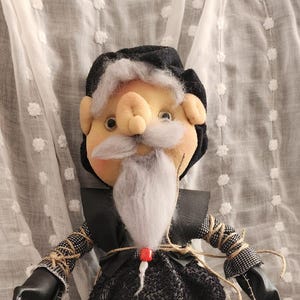 May include: A handmade doll with a whimsical design. The doll features a light skin tone face with a white beard and black hair. It wears a black outfit with black boots and has a small red bead. A heart-shaped rug is in the foreground.