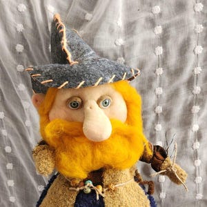May include: A handmade doll with a whimsical design. It features a gray hat, a large orange beard, and a tan and blue outfit. The doll has a long nose and is holding a small twig. The doll's feet are wearing plaid and blue shoes.