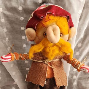 May include: A handmade doll with a yellow face, orange beard, and a red hat. The doll wears a brown vest and pants, with striped arms and small black shoes. An orange crocheted pumpkin sits nearby.