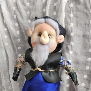 May include: A handmade doll with a whimsical design. It features a light skin tone, a large nose, and a white beard. The doll wears a black vest, blue pants, and black shoes. The arms are wrapped in fabric.