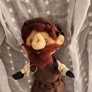 May include: A handmade doll of a bearded man wearing a brown outfit with a plaid scarf and belt. The doll has a large nose and a red mustache. A small figurine is next to the doll.
