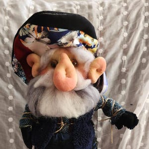 May include: A handmade gnome doll with a large pink nose, gray beard, and a black hat with a floral pattern. The gnome wears a blue vest and pants, with black shoes. The doll is on a white dotted fabric.