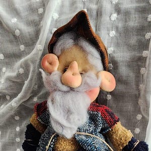 May include: A handmade gnome doll with a white beard, pink nose, and ears, wearing a plaid outfit in brown, blue, and red. The doll has black shoes and is standing near a small pumpkin and two red ladybugs.