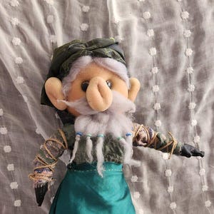 May include: A handmade doll of a gnome with a large nose, white beard, and green outfit. The doll has a green hat, a teal apron, and black shoes. The arms are wrapped in twine and fabric.