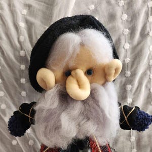 May include: Handmade gnome doll with a black hat, white beard, and a red and black plaid vest. The doll has blue eyes and is wearing black pants and blue shoes. The doll is made of fabric and yarn.