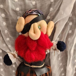 May include: A handmade doll with a red beard, plaid pants, and a black leather blindfold. The doll has tan ears and a black leather belt. A small, colorful, round trinket and a small crocheted figure are also present.
