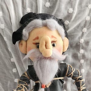 May include: A handmade fabric doll depicting an elderly man. The doll features a large nose, white beard, and a black hat. The doll is dressed in a black and gray outfit with a gray skirt and black shoes. The doll is approximately 12 inches tall.