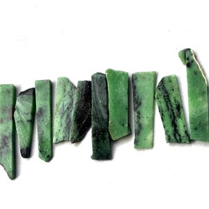 May include: A collection of rectangular green stones with dark green and black veining. The stones are arranged on a white surface, showcasing their natural textures and variations in color. These stones could be used for jewelry making or other crafts.