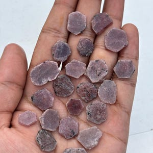 Puede incluir: Una colección de gemas hexagonales en bruto, en tonos rosados y púrpuras. Las piedras se sostienen en una mano, mostrando sus superficies naturales y sin pulir. Algunas piedras tienen inclusiones oscuras, lo que añade a su carácter único. Las gemas son un producto natural.